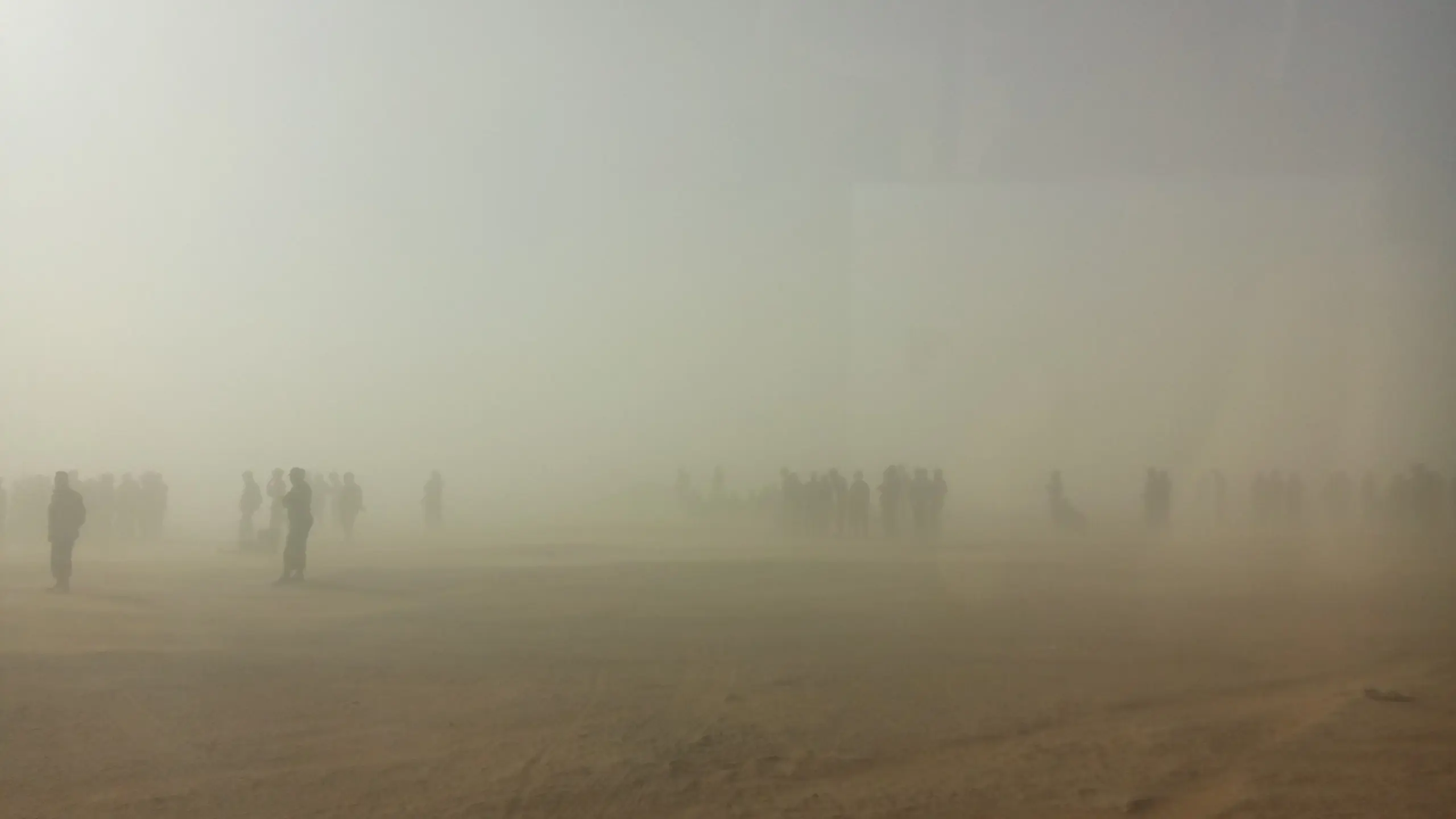 Photo of a dust storm. Very bury image with a yellow/brown haze covering the sky with groups of people just recognizable in the background