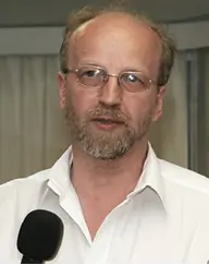 Portrait photo of a man in a white shirt with a beard and glasses talking into a microphone