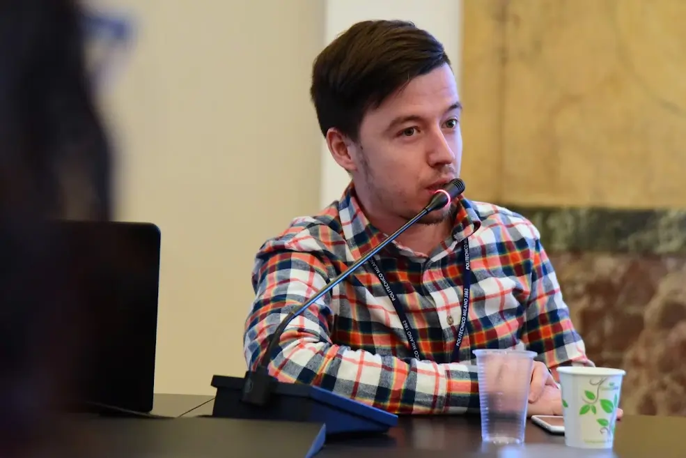 Photo of a brown-haired man in his early thirties wearing a check shirt talking into a microphone