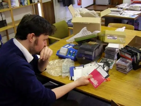 Man sitting at a desk looking intensely at a selection of old floppy disks, memory cards, tapes, and CDs