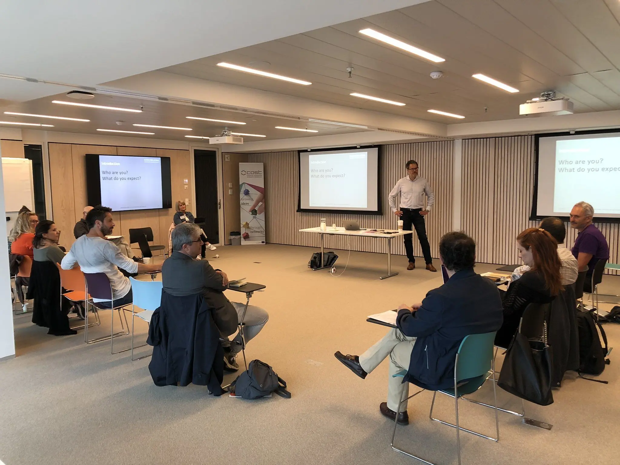 A man standing in a room presenting to a group of people sat on chairs in a U configuration