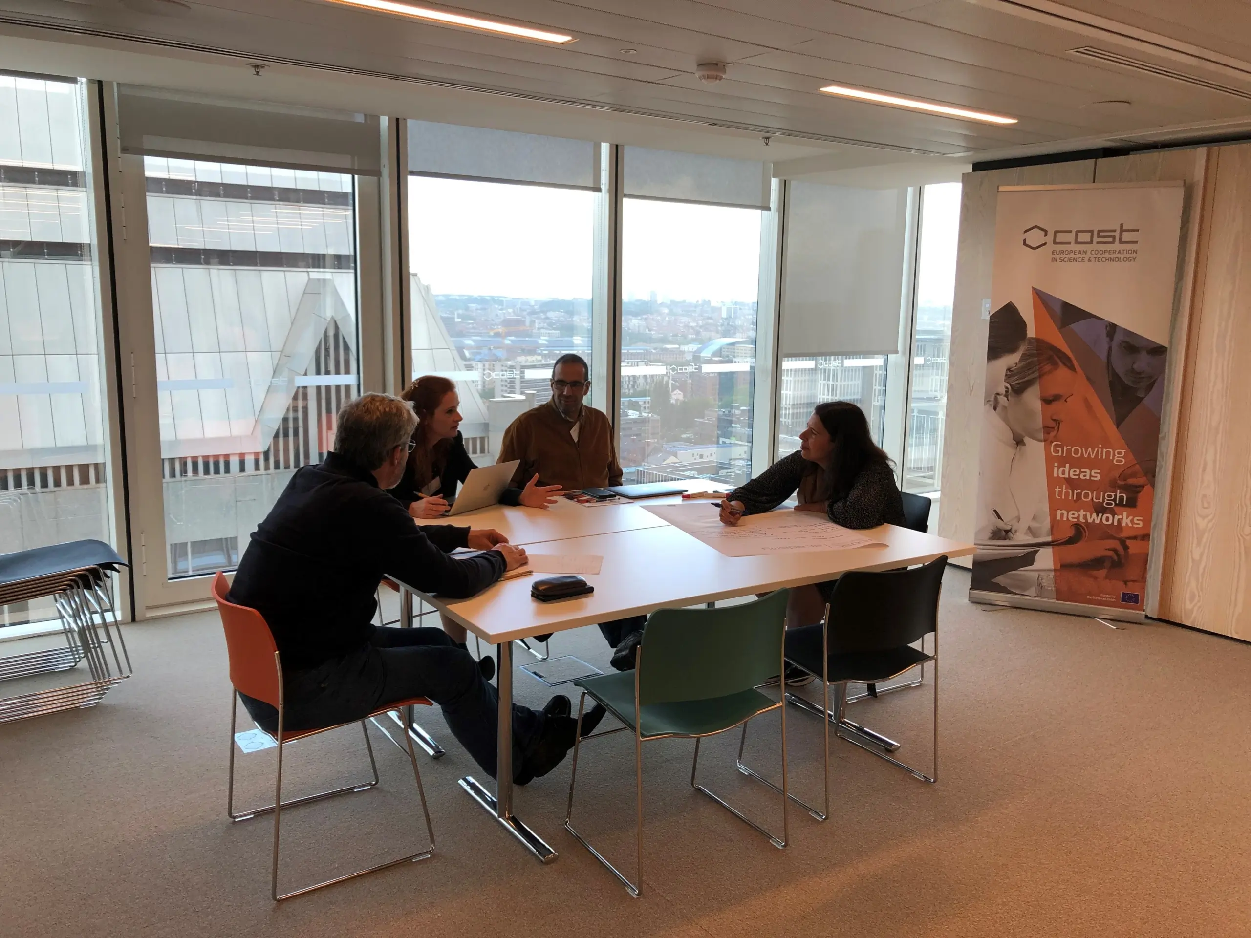 Four people sat at a square table in a large office in discussion