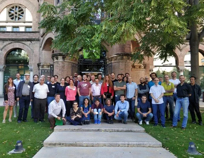 Group picture of 60+ researchers standing together outside a building with a large tree in Granada