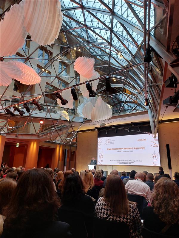 Photo taken during a conference showing a packed audience listing to a male keynote speaker on a podium. Above the audience is an architecturally interesting glass and metal ceiling with suspended white sculptures.