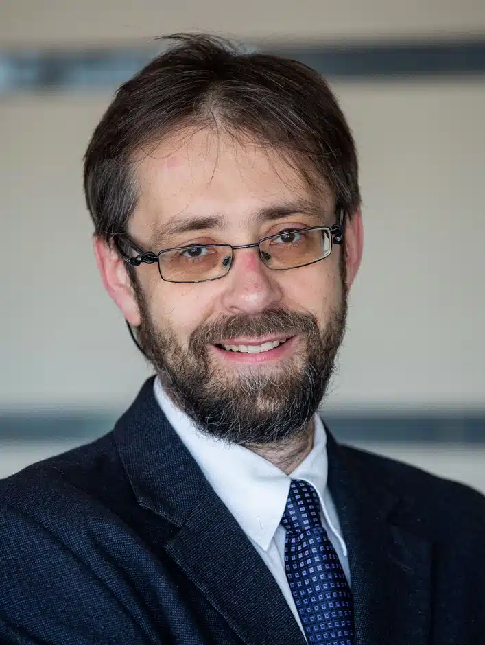 Portrait photo of a middle aged man wearing square black glasses. He has short black hair, and a black moustache and beard