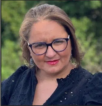 Portrait photo of a woman with shoulder length blond hair. She is smiling to camera and dressed in a black top, pink lipstick, and large framed black glasses.