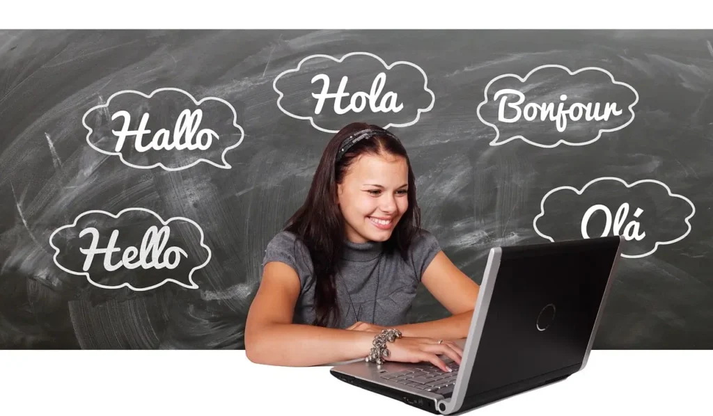 Girl sat infront of a computer with the words "Hola; Bonjour; Ola; Hello" on a blackboard behind her  