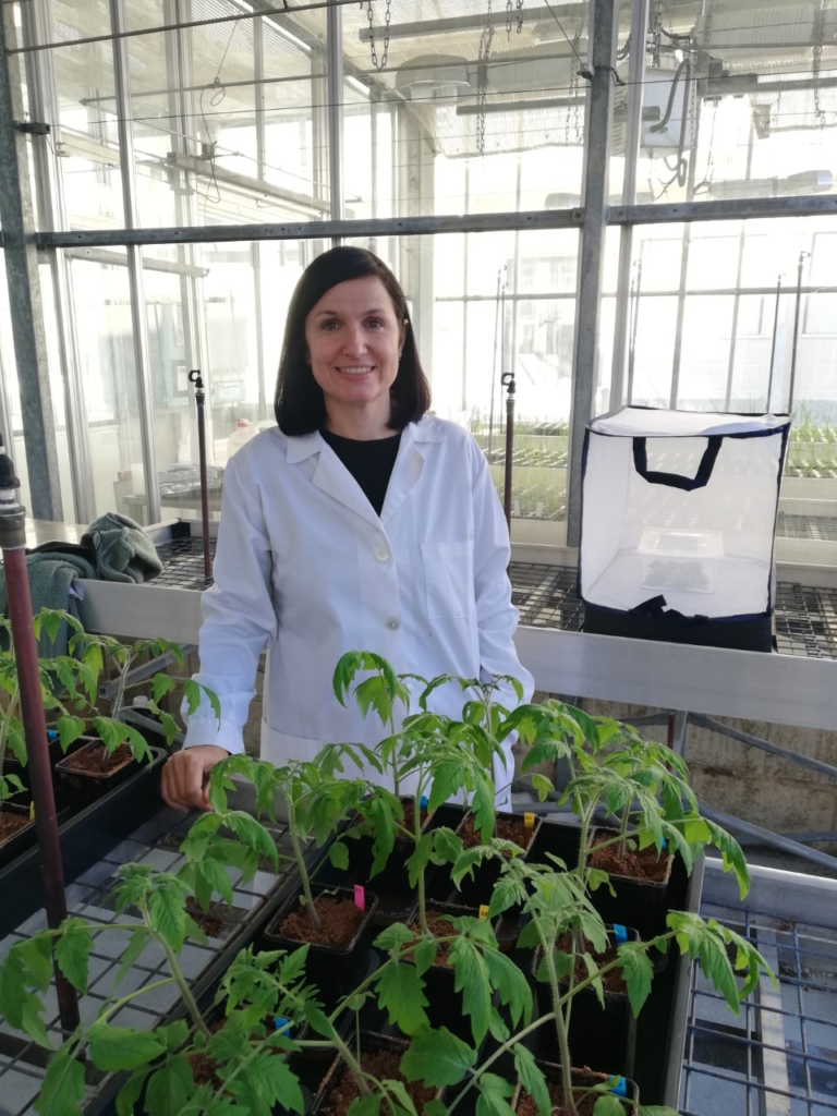 A young woman in a white lab coat is standing in a greenhouse.