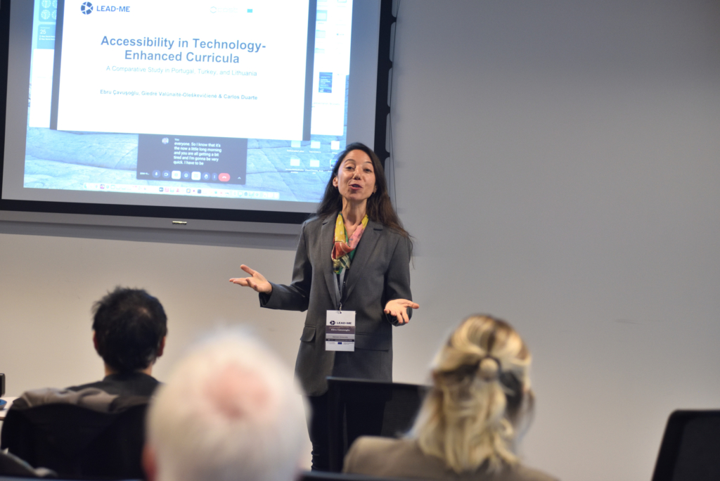 Middle-aged woman with long dark hair wearing a grey blazer and rainbow neck scarf presenting at the LEAD-ME final event. A screen behind her shows a slide with the text "Accessibility in technology: enhanced curricula"