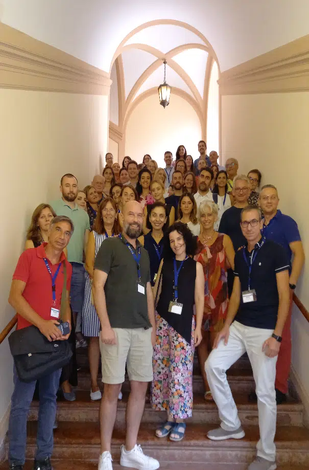 A group photo of people standing on a staircase in what appears to be a historic building, with distinctive vaulted arches and a hanging lantern overhead. Several people wearing name badges are positioned in the front.