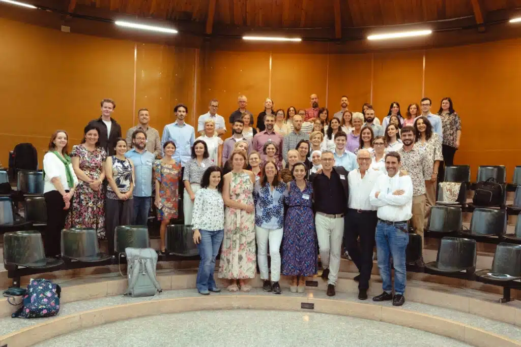 A large group photo of people standing in what appears to be a tiered lecture hall or auditorium with wooden walls and stadium-style seating.