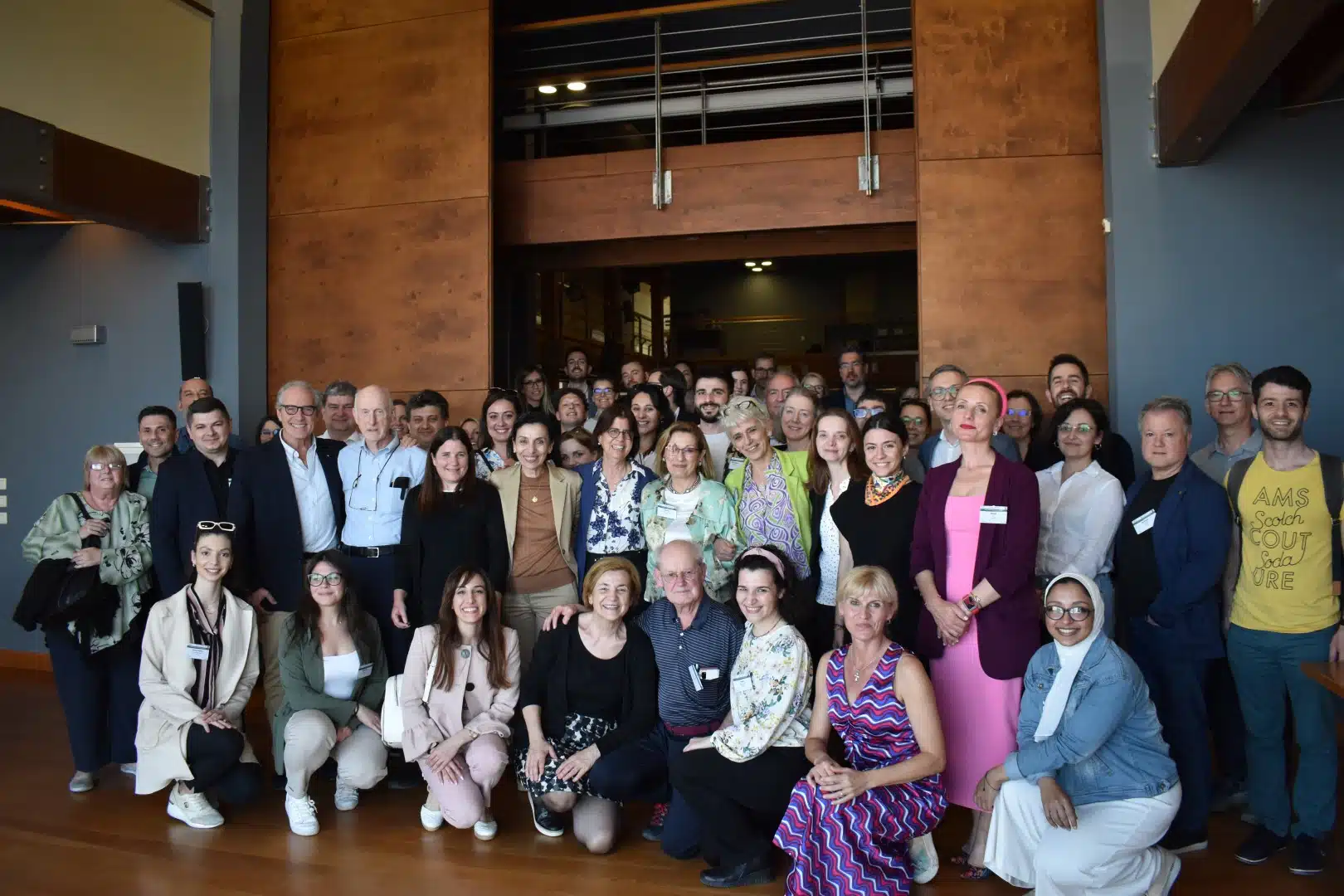 A large group photo of approximately 40-50 people in casual business attire, taken in what appears to be a modern building lobby or conference space with wooden architectural features and gray walls.