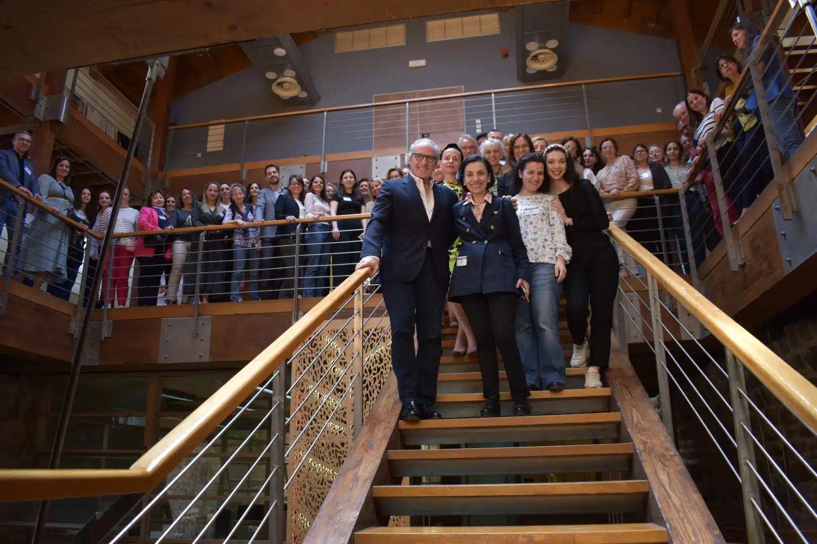 A large group photo taken on and around a modern wooden staircase with metal railings. Several people stand on the stairs in the foreground while many others are gathered on the landing above.