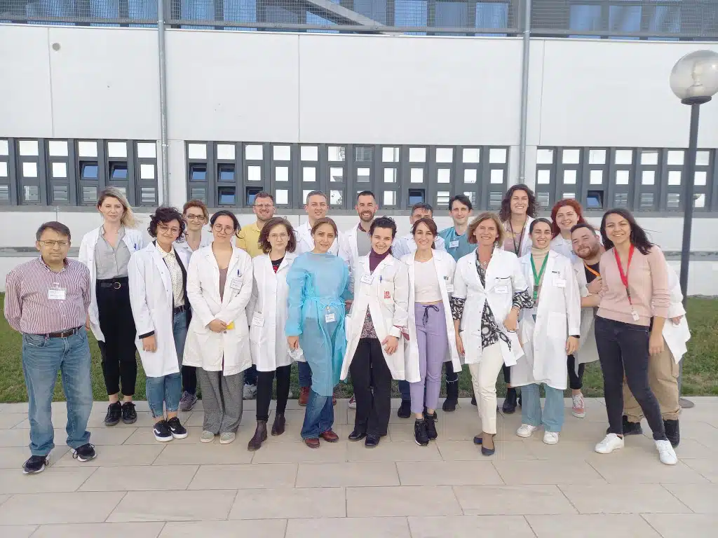 A group photo of approximately 15-20 people, most wearing white lab coats or medical attire, standing outside what appears to be a medical or research facility. One person in the center is wearing light blue scrubs.