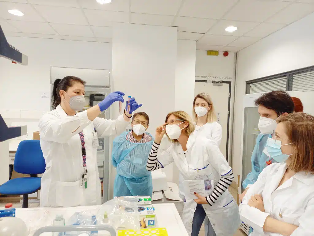 The image shows people wearing masks and white lab coats working at a laboratory bench with scientific equipment. A person in the foreground is conducting some kind of lab procedure.