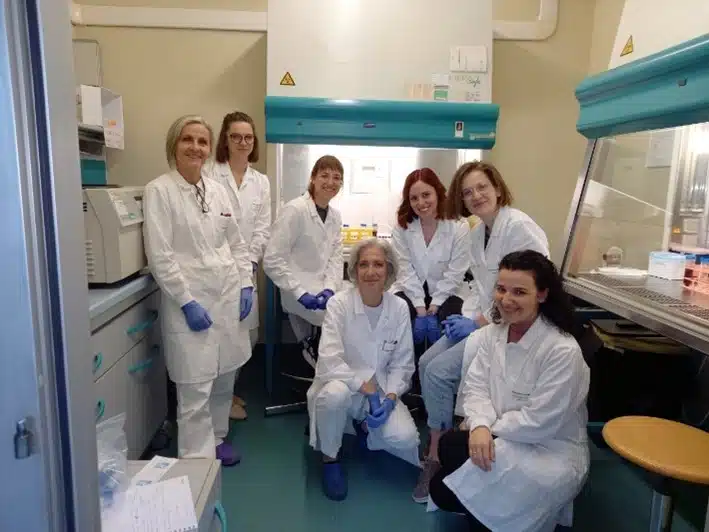 A group of seven people in white lab coats and blue gloves posing inside a laboratory setting, with lab equipment and safety hoods visible in the background.