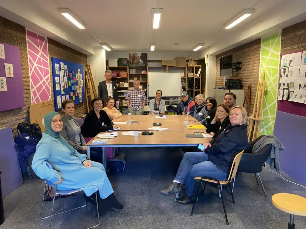 A multi-faith  and multi-age group of  men and women meet to represent migrants. 12 people sit round a large square table in a workshop