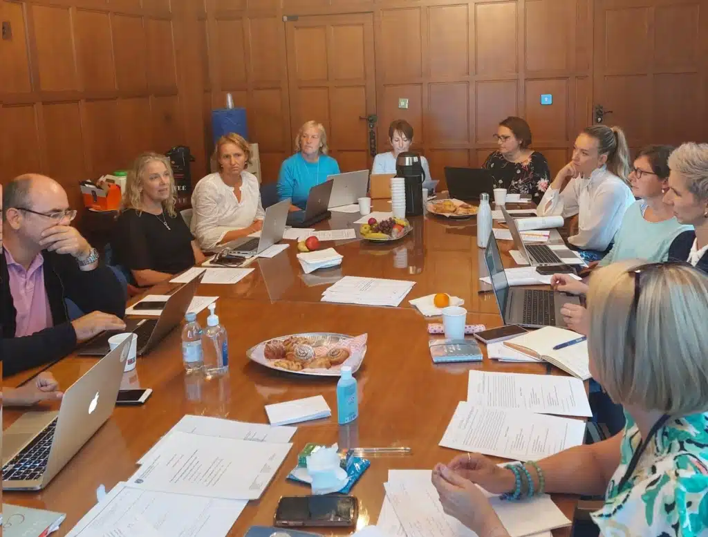 The image shows a business meeting or workshop taking place around a large wooden conference table in a room with wooden paneled walls. About 10-12 people are seated around the table, most with laptops open in front of them. The attendees appear to be predominantly women with one or two men visible.