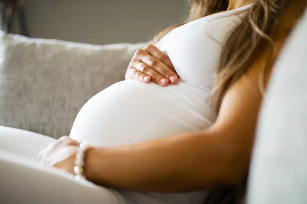 The image shows a pregnant woman sitting on a light-colored couch. She's wearing a white fitted top and has her hand gently resting on her rounded belly. Only the midsection and part of her arm are visible, along with some blonde hair. She's wearing a bracelet on her wrist. The photo has soft lighting and focuses on the maternal gesture of cradling her pregnancy.