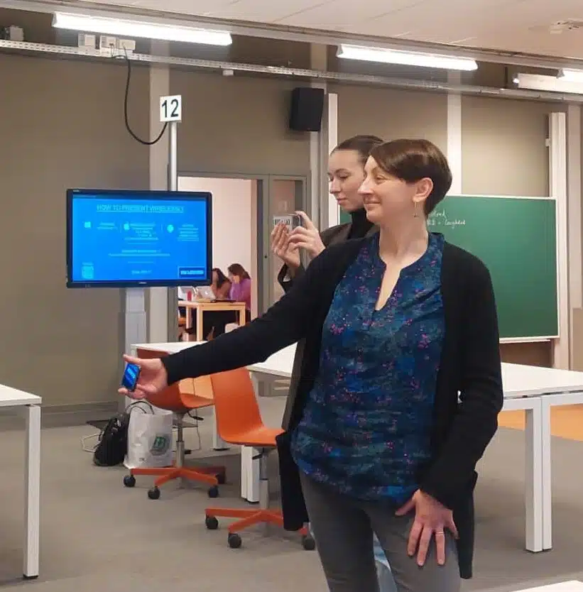 The image shows a classroom or training room setting. In the foreground, a person with short dark hair wearing a blue patterned top and black cardigan is standing and holding what appears to be a small device or remote in their hand. Behind them, another person is holding up what looks like a smartphone or camera.