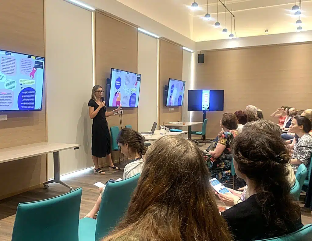 The image shows a presentation or workshop in a modern meeting room. A person wearing a black dress and glasses is standing at the front, speaking into a microphone. They appear to be presenting information shown on multiple display screens mounted on the light wood-paneled walls.